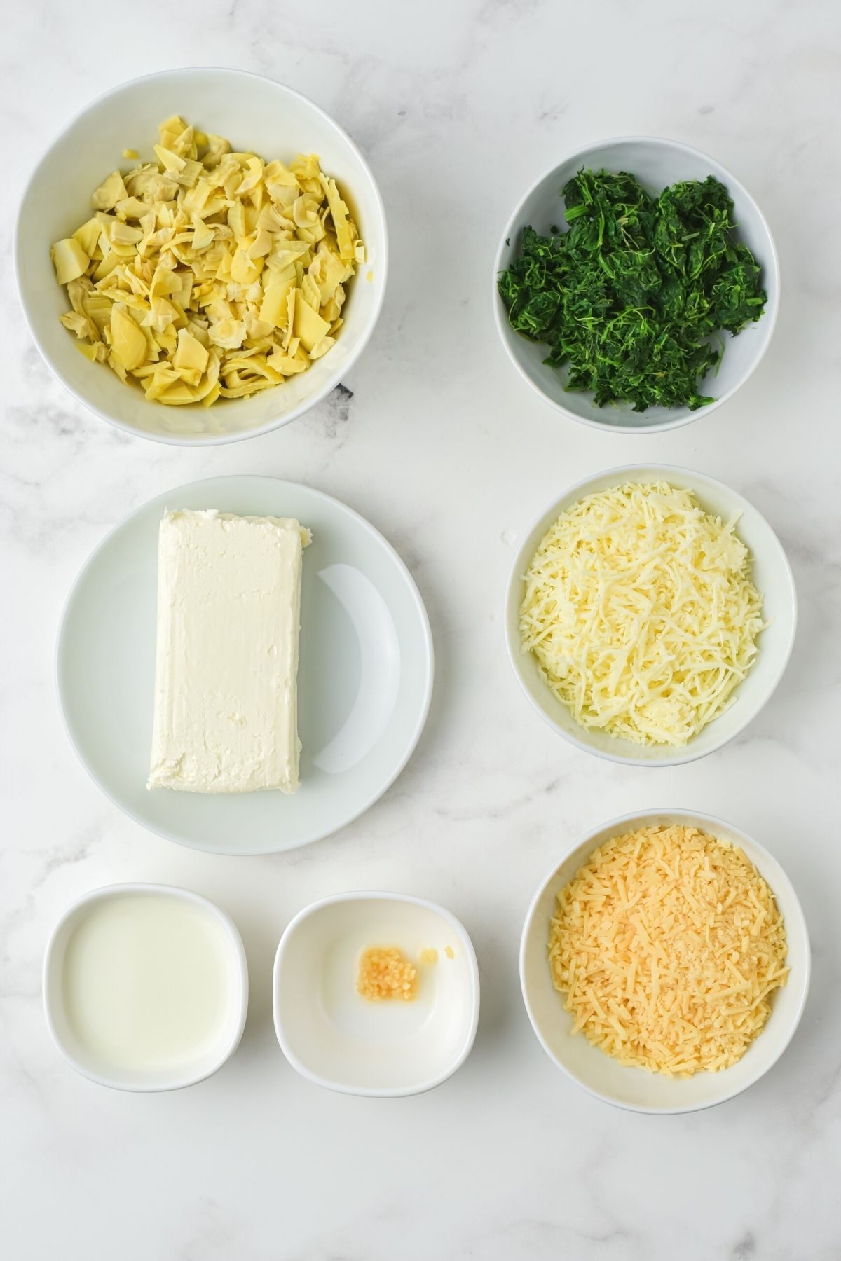 ingredients in white bowls on counter: drained artichokes, frozen spinach, block of cream cheese, shredded mozzarella, parmesan cheese, milk, garlic