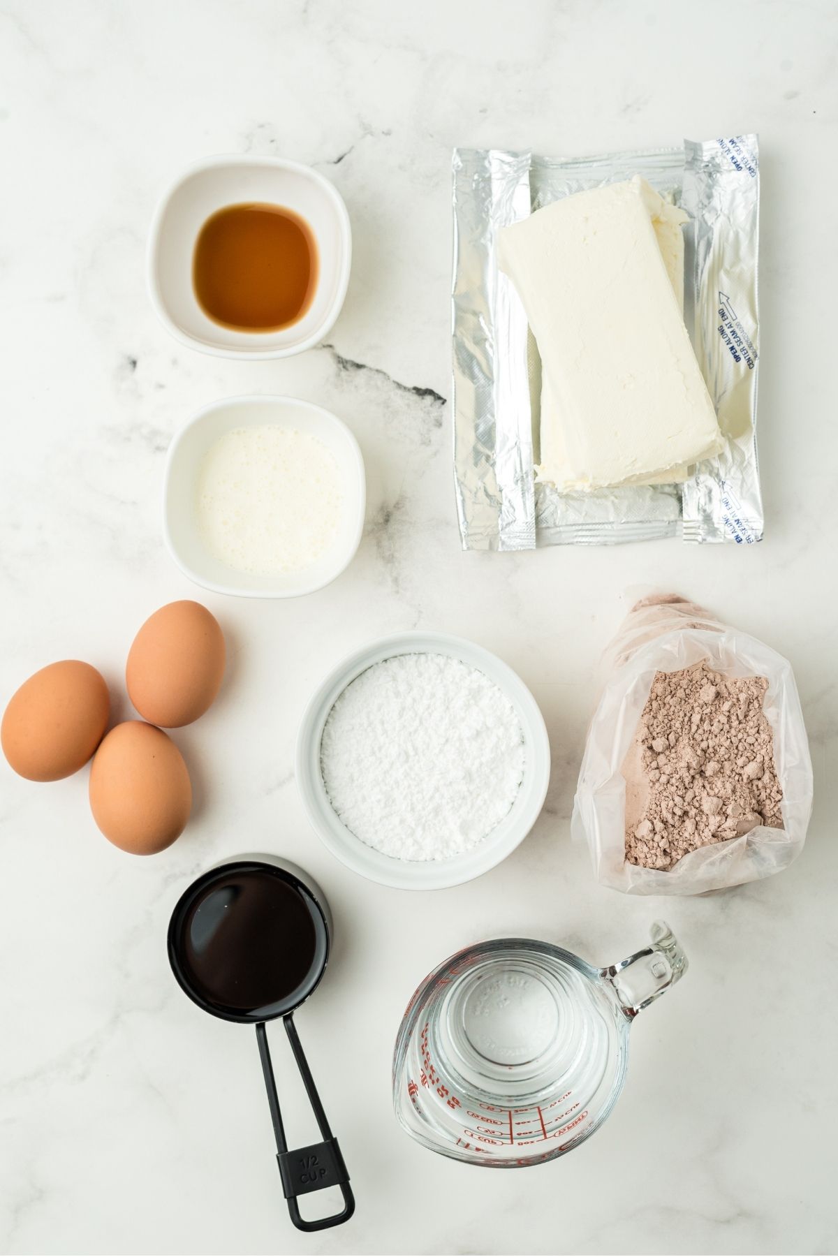 ingredients on white counter: vanilla, two blocks cream cheese, red velvet cake mix, powdered sugar, 3 eggs, heavy cream, vanilla