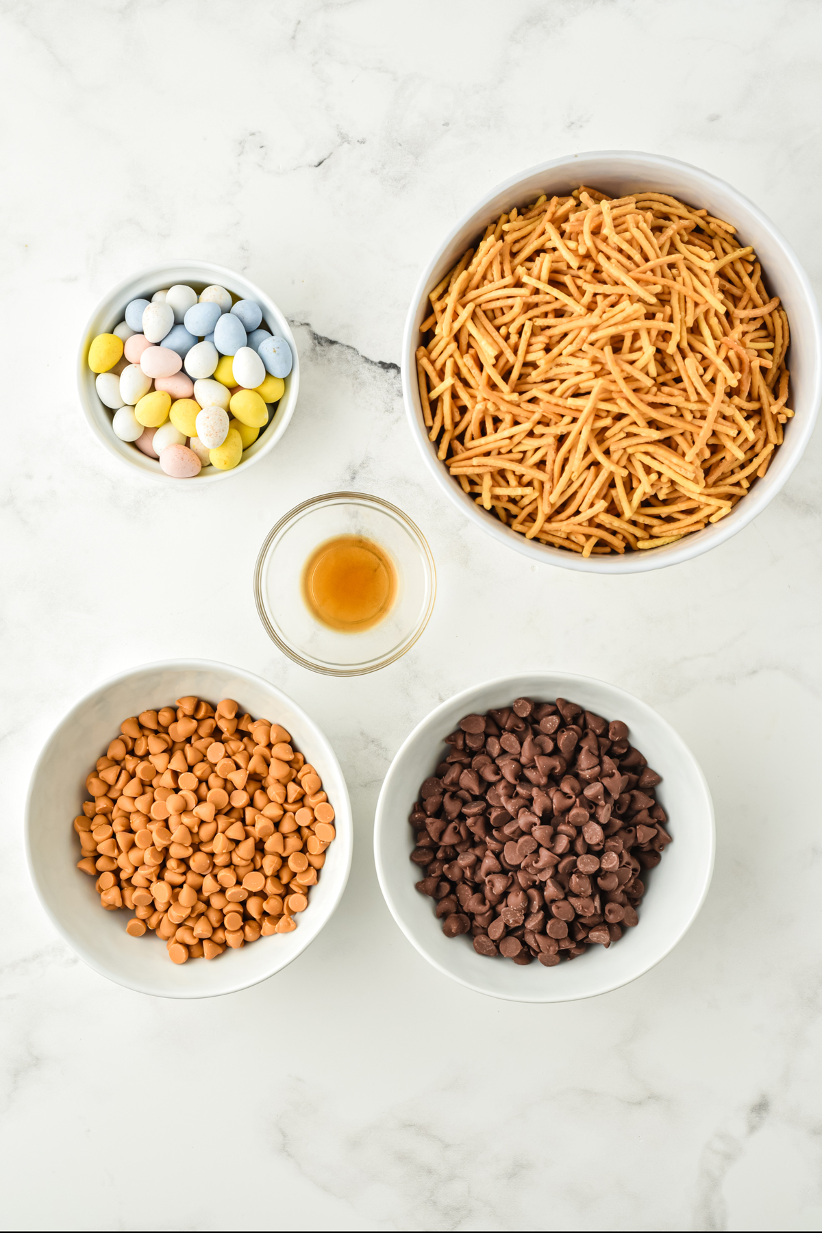 Ingredients of bird's nest Cookies on a counter. These are milk chocolate chips, butterscotch chips, Chow Mein noodles, vanilla extract, and egg candies. 
