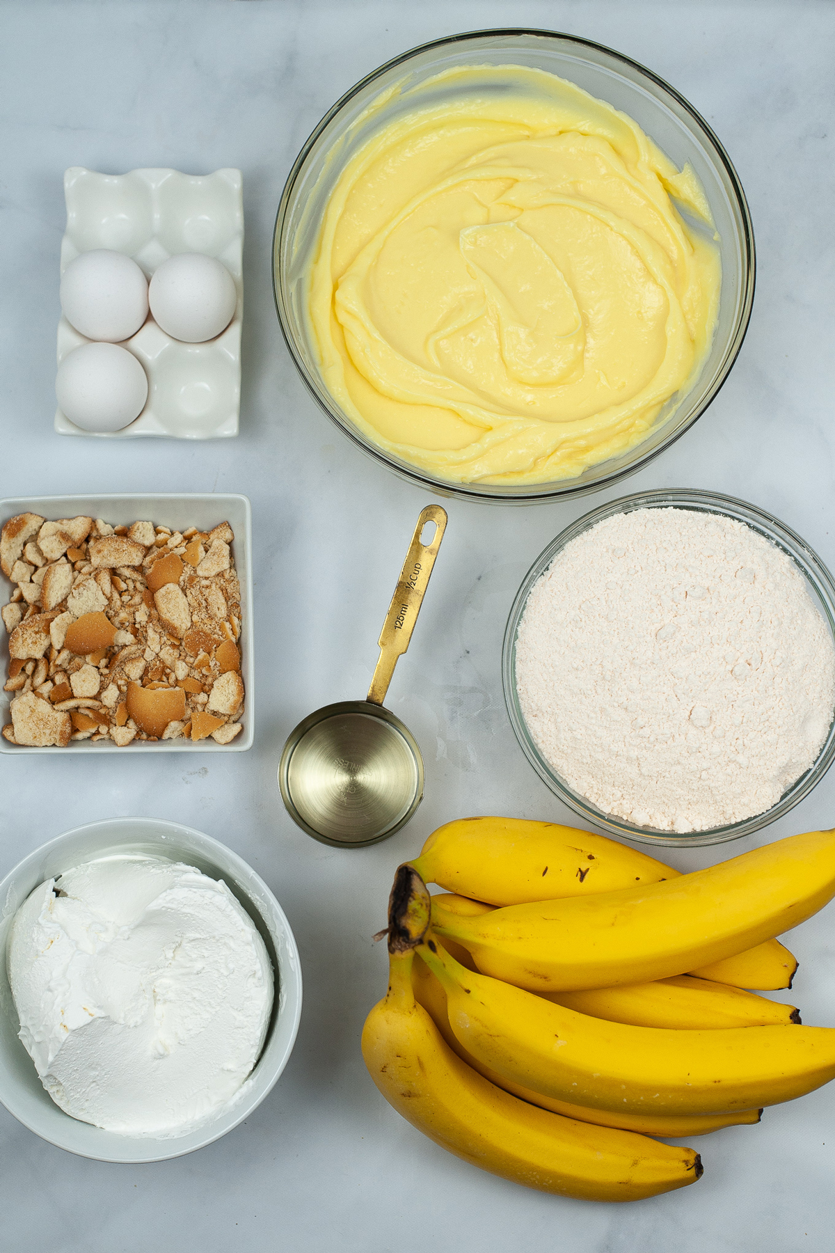 Ingredients of Banana Pudding Poke Cake on a white counter. The ingredients are yellow cake mix, eggs, bananas, water, oil, banana pudding, and vanilla wafer crumbs. 