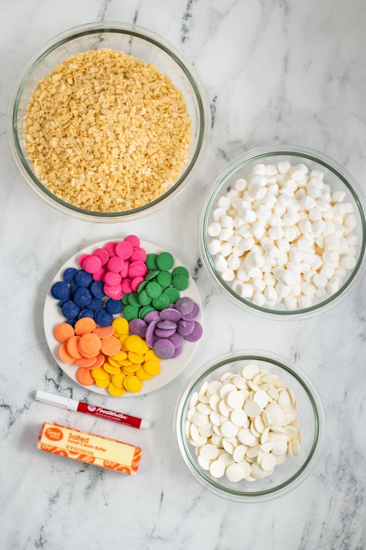 ingredients on white counter: colorful candy melts, mini marshmallows, edible ink pen, butter, bowl of rice Krispie cereal