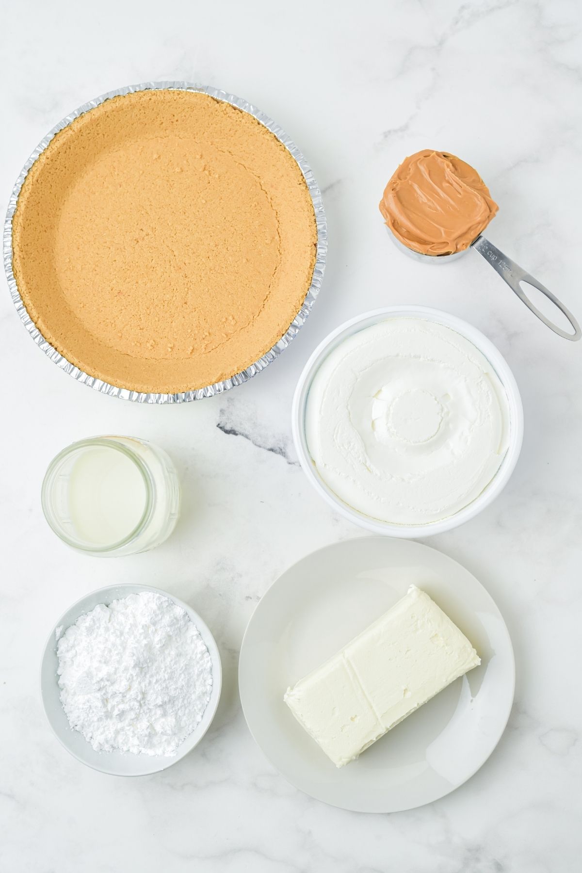 ingredients on white counter: graham cracker pie crust, peanut butter, Cool Whip, block of cream cheese, powdered sugar, milk