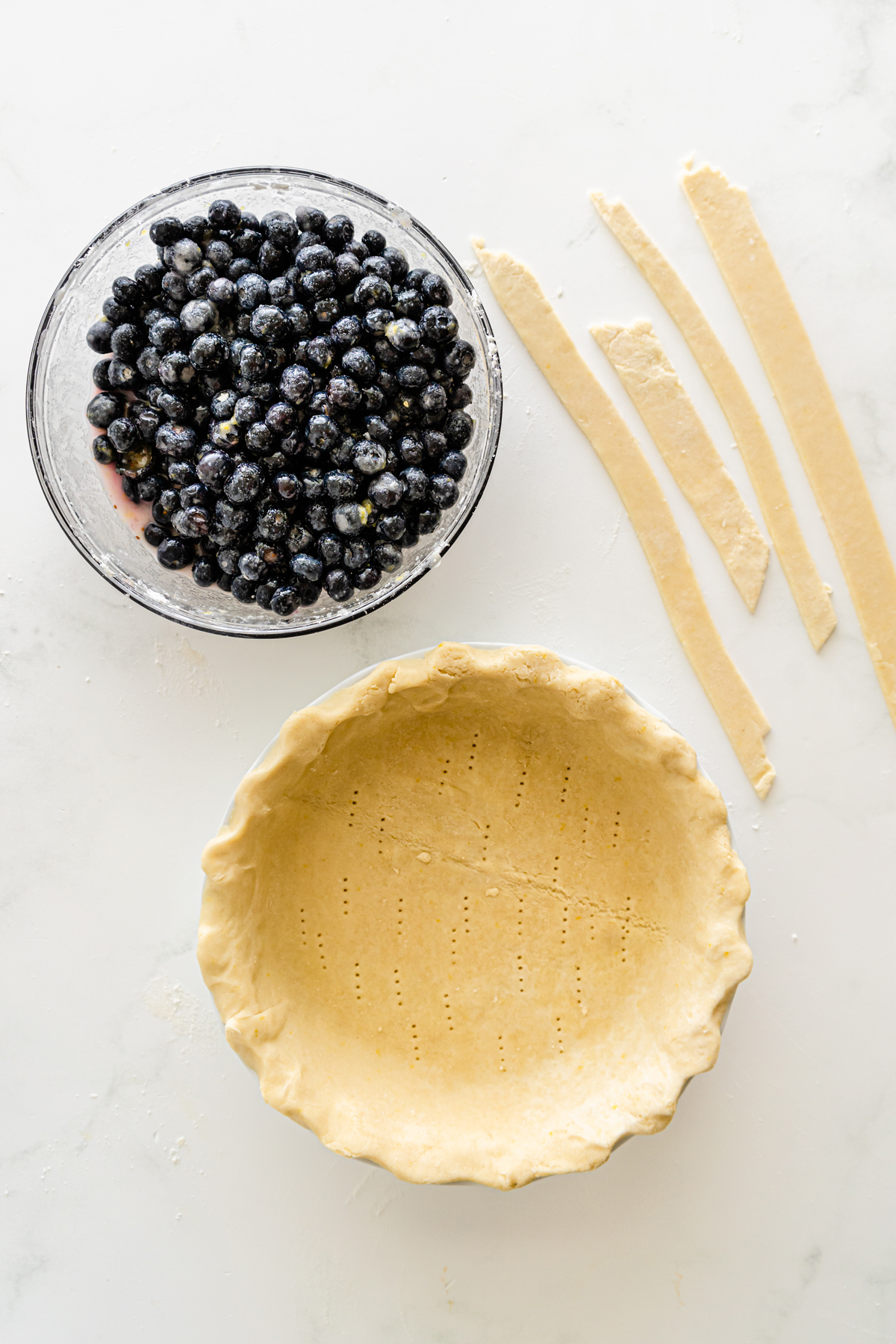 Top view of a table with a pie dish with round pastry on bottom, a big bowl of blueberries next to it, with strips of pastry next to it. 