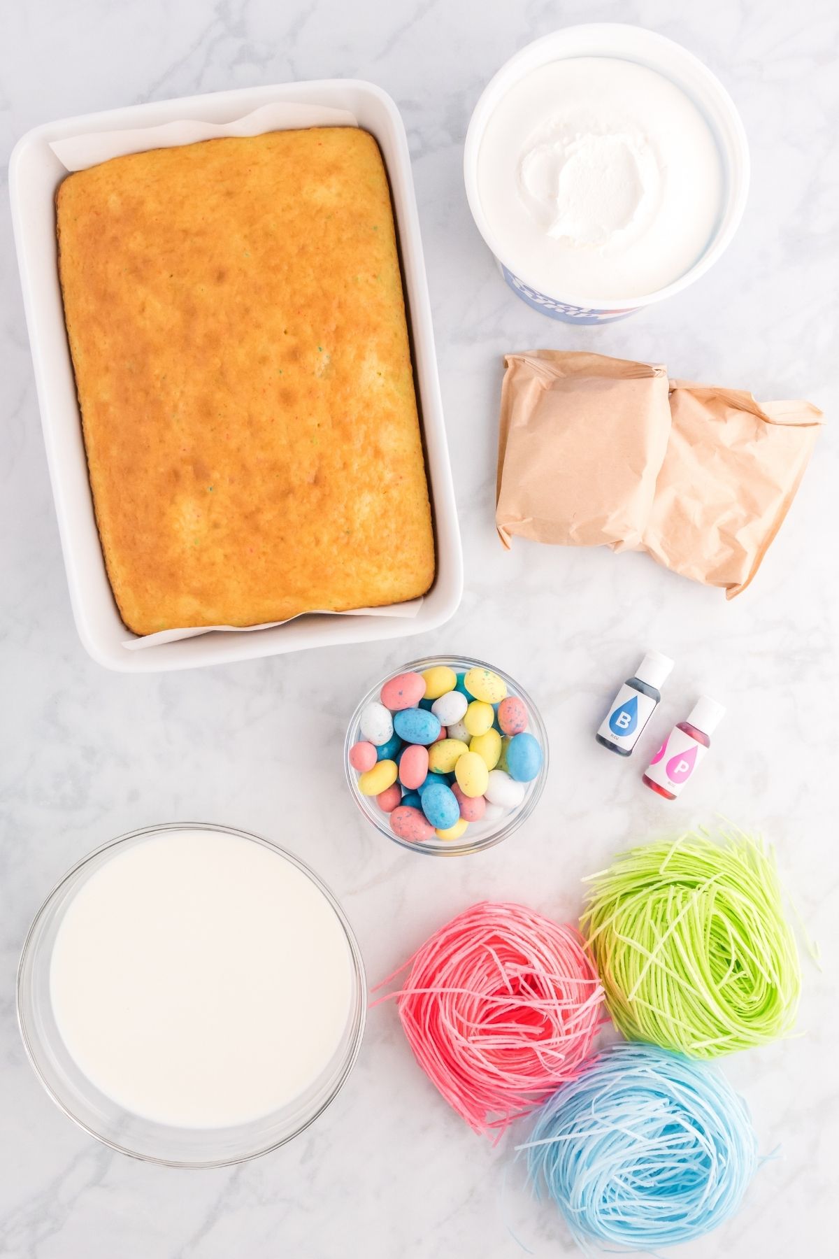 ingredients on white counter: baked rainbow cake, pudding packets, milk, food coloring, edible candy grass, Easter egg candies