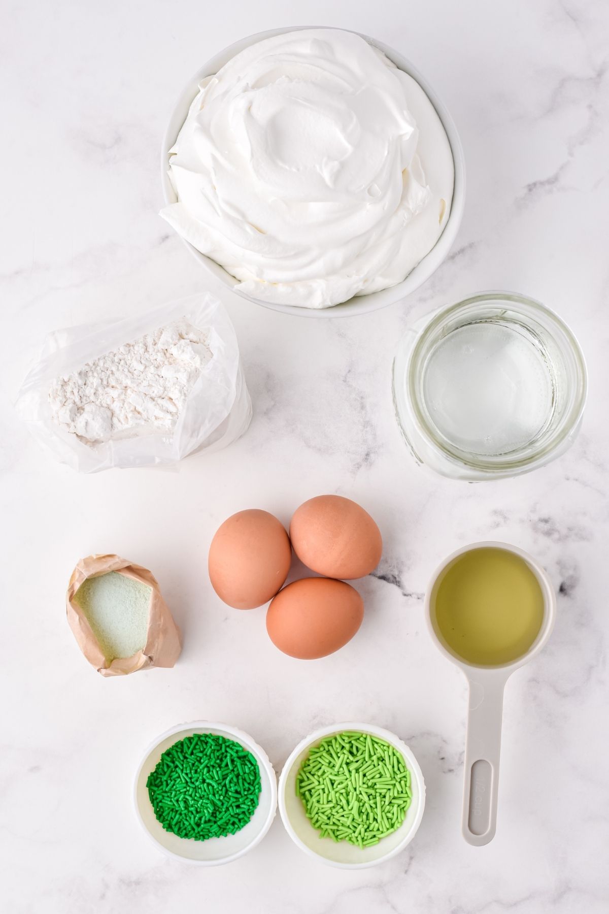 ingredients on white counter: whipped topping, water, oil, eggs, cake mix, jello, green sprinkles