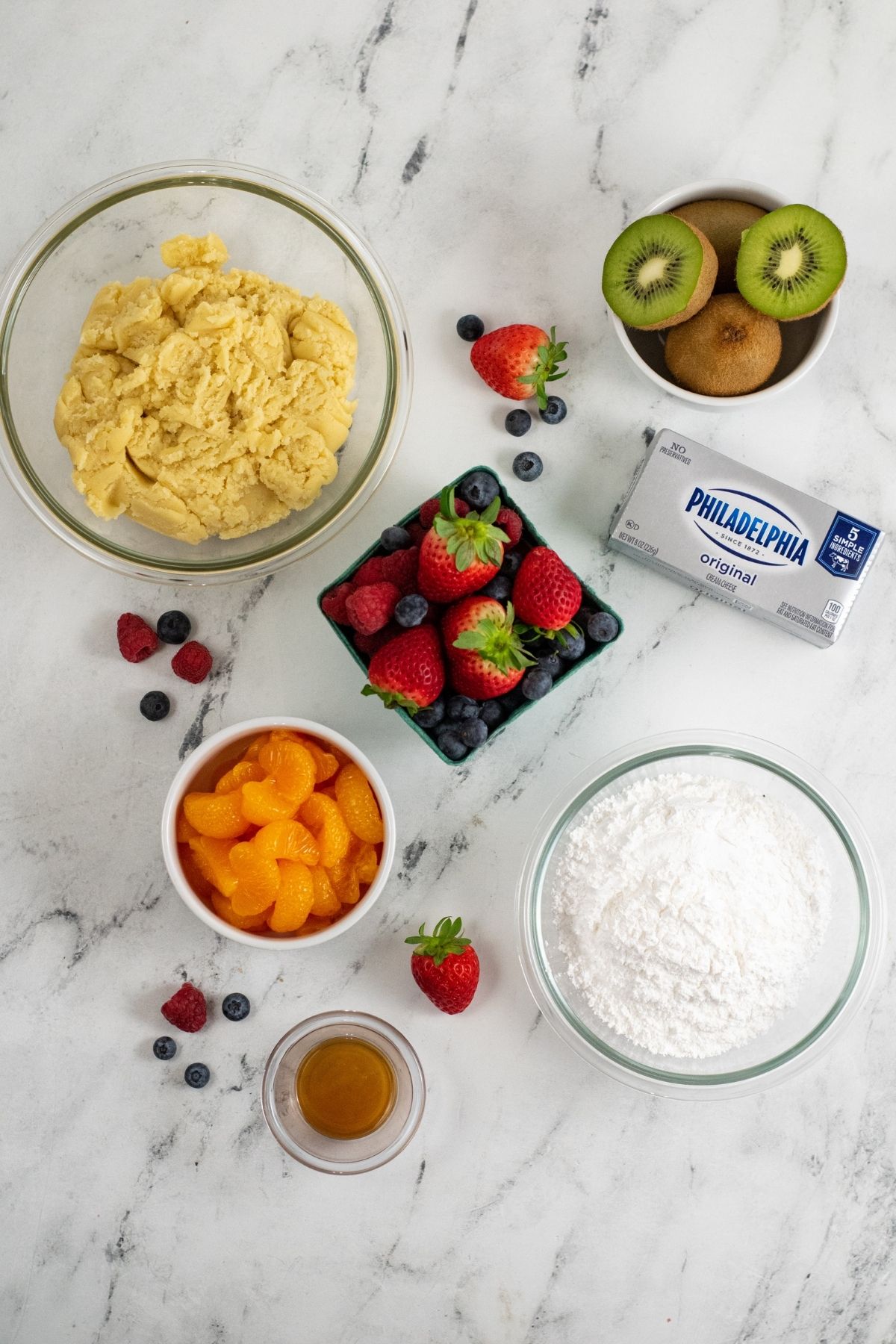 ingredients on white counter: cookie dough, cream cheese, powdered sugar, strawberries, blueberries, kiwi, and mandarin oranges