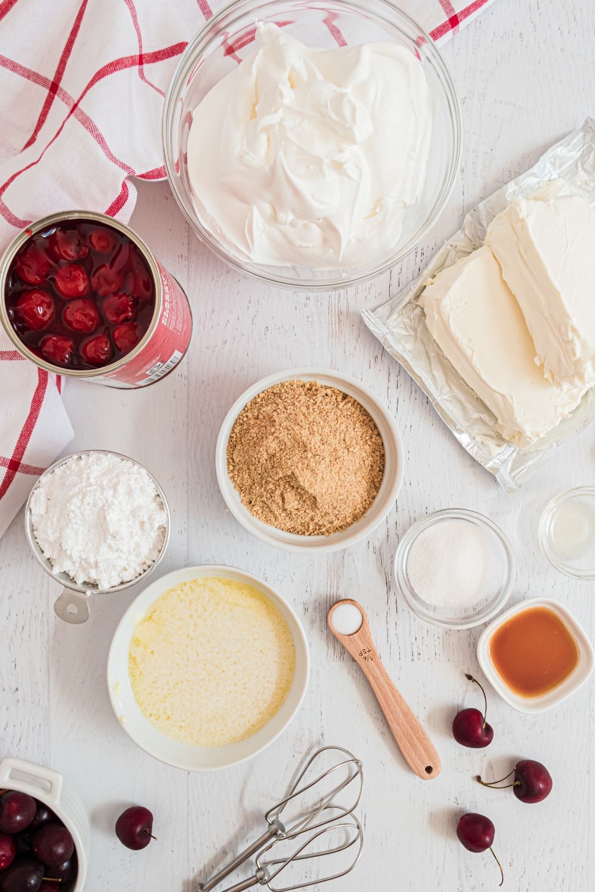 ingredients on white wooden counter: crushed graham crackers, whipped topping, two blocks cream cheese, can of cherry pie filling, powdered sugar, melted butter, vanilla, lemon juice, salt, and sugar