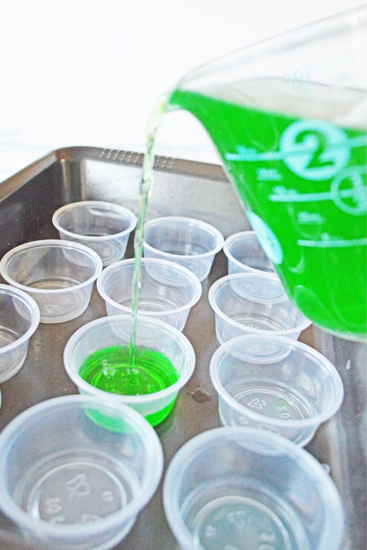 measuring cup with green jello being poured into plastic shot cups