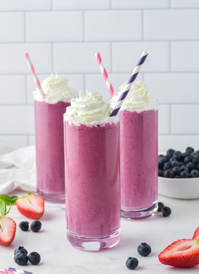 Three glasses filled with pink smoothie mix, with stripy straws in them and chopped berries on the work top.