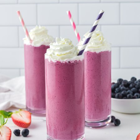 Three glasses filled with pink smoothie mix, with stripy straws in them and chopped berries on the work top.