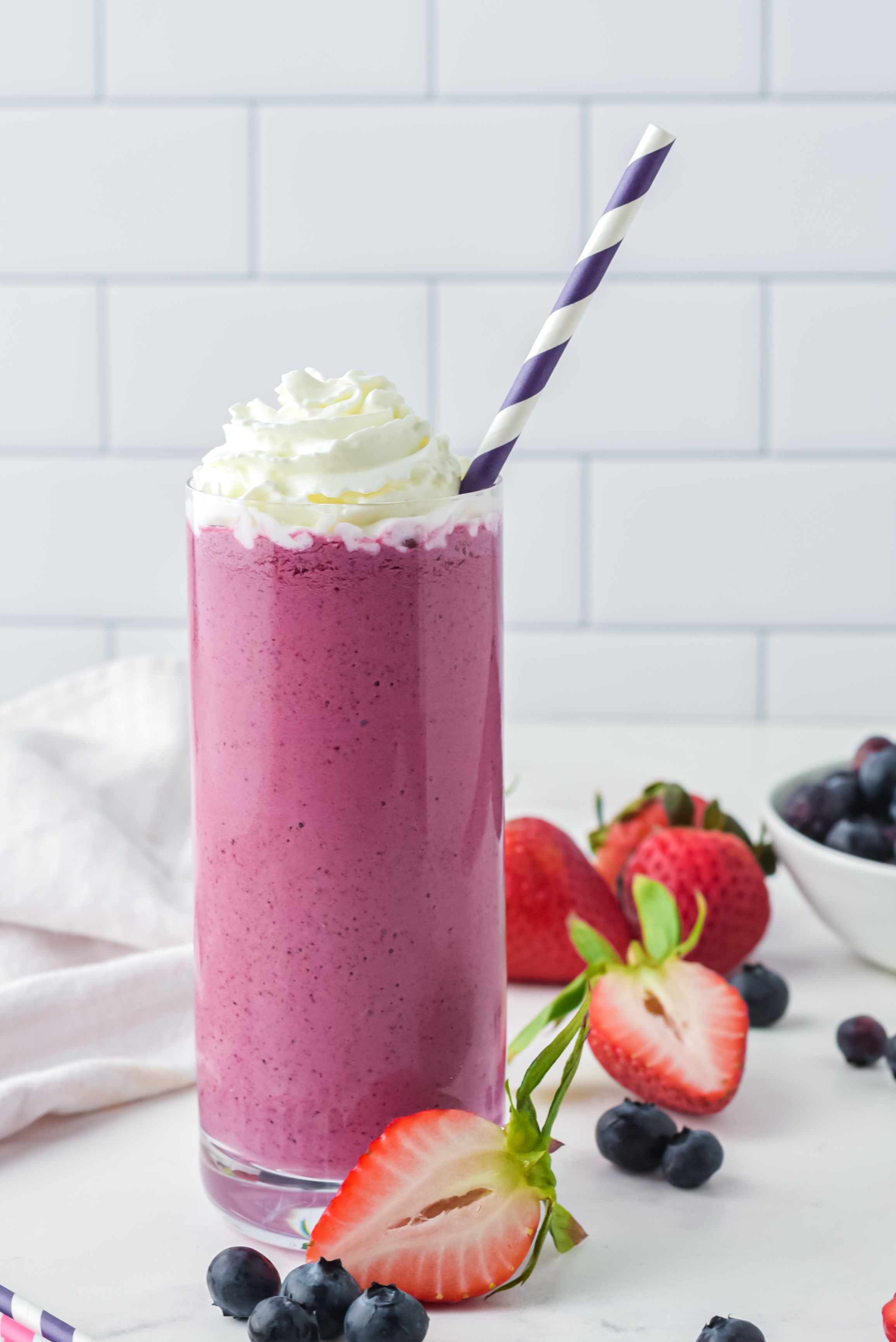 Glass filled with pink berry smoothie mix, with whipped cream on top and a purple stripy straw in the top, surrounded by chopped strawberries and blueberries on the table around it. 