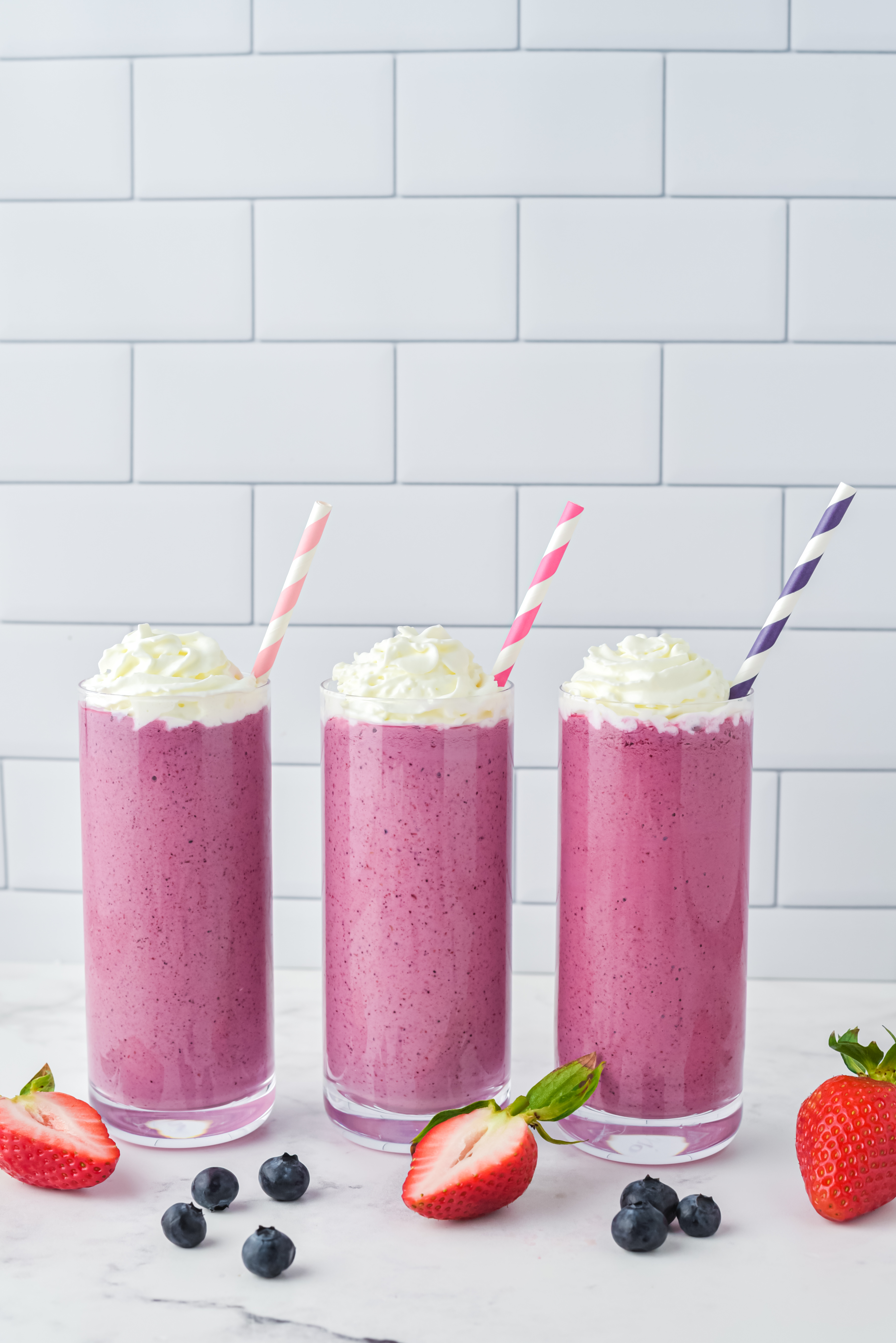 Three glasses with pink berry smoothie mix in them, stripy straws in the top, and chopped berries on the white table around the glasses.