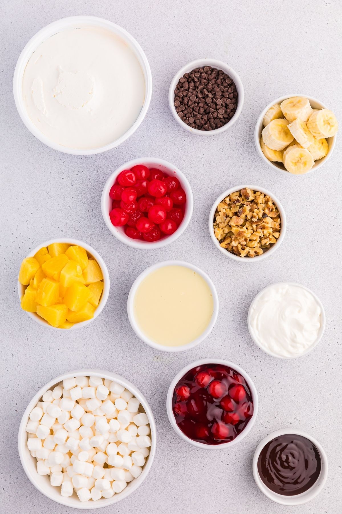 ingredients in bowls on white counter: Cool Whip, mini chocolate chips, sliced bananas, chopped walnuts, maraschino cherries, pineapple, condensed milk, sour cream, cherry pie filling, mini marshmallows, chocolate sauce