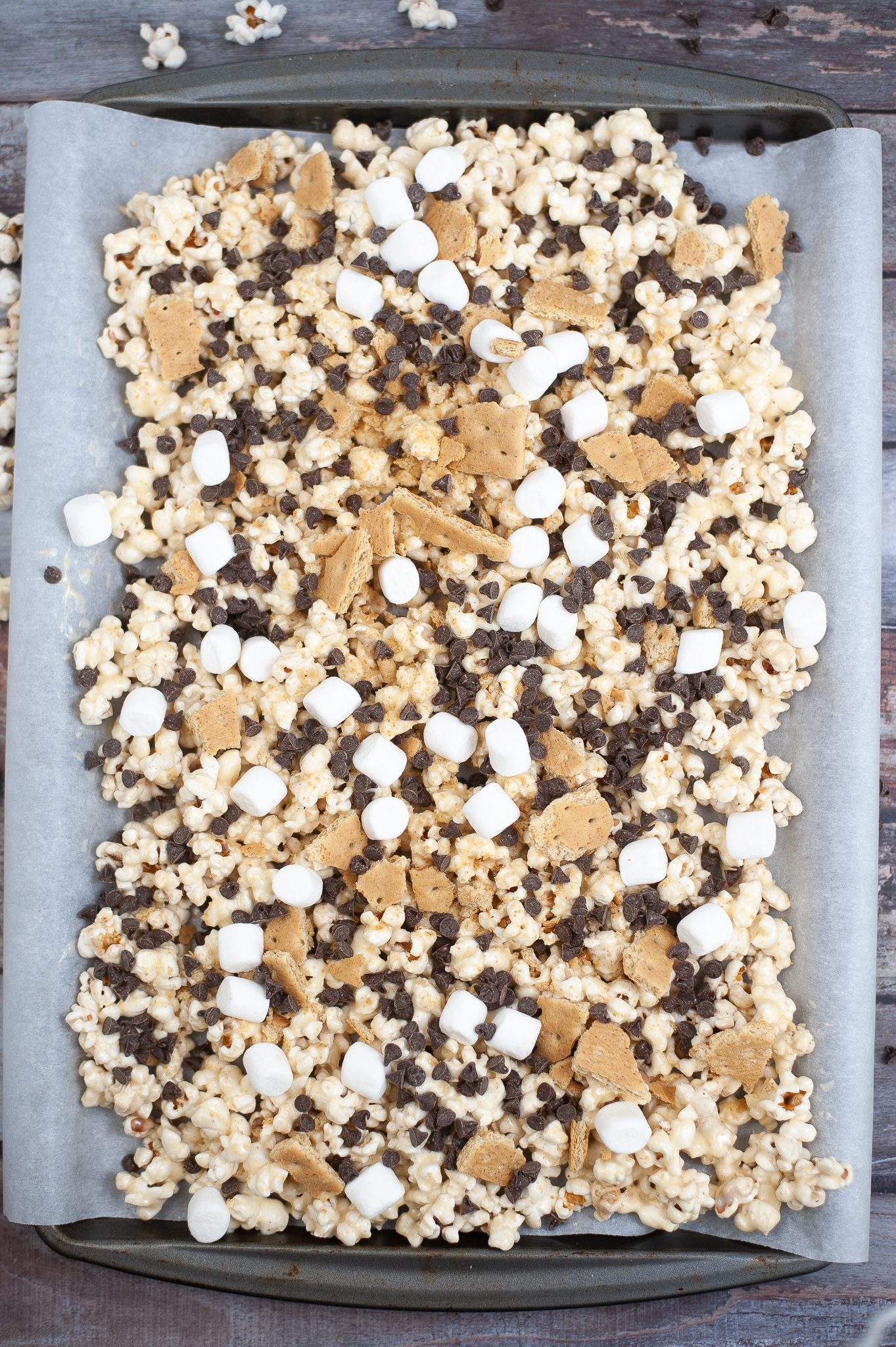 Popcorn on a tray with marshmallows, crushed graham crackers, and chocolate chips on top on a baking tray with parchment paper.