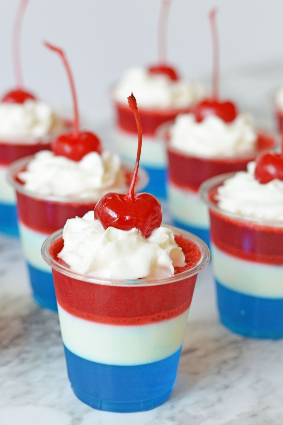 red, white and blue jello shots in small cups with whipped cream and maraschino cherries