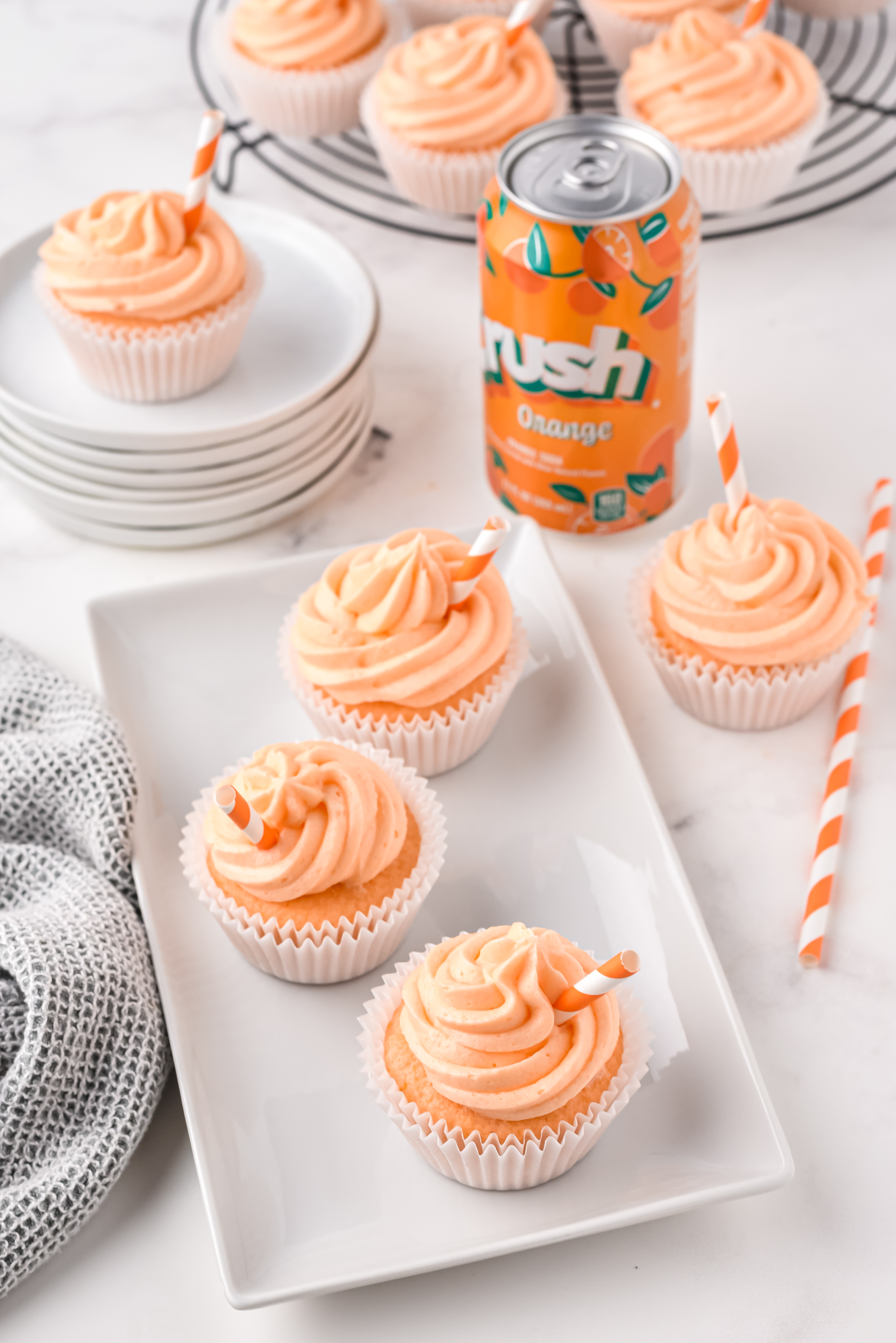 Cupcakes with orange frosting and mini stripy straws in them on a white rectangle plate, with a can of orange crush soda next to it and other orange crush cupcakes on the table with it.