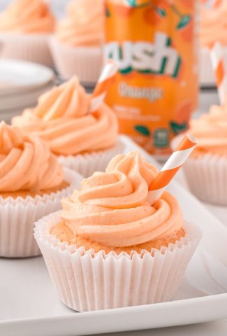 Cupcake with orange icing with short stripy straw in it, and a can of orange crush soda behind it and other cupcakes on the table too.