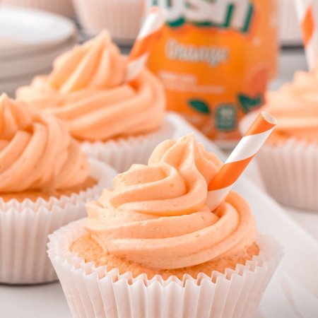 Cupcake with orange icing with short stripy straw in it, and a can of orange crush soda behind it and other cupcakes on the table too.