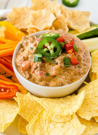 Close up of Crockpot Rotel Dip in a white bowl, on a plate with sliced pepper, corn tortilla chips, and sliced cucumber on it.