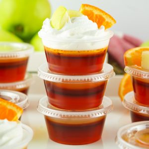 A stack of three sangria jello shots in small plastic cups, the top one is garnished with whipped cream and small orange and apple slices.