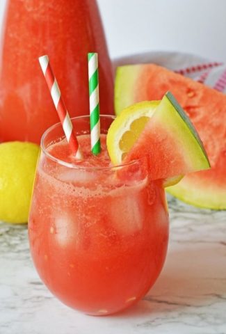 A hurricane glass on a table filled with red watermelon lemonade with two stripy straws in it with slices of watermelon and lemon on the rim of the glass.