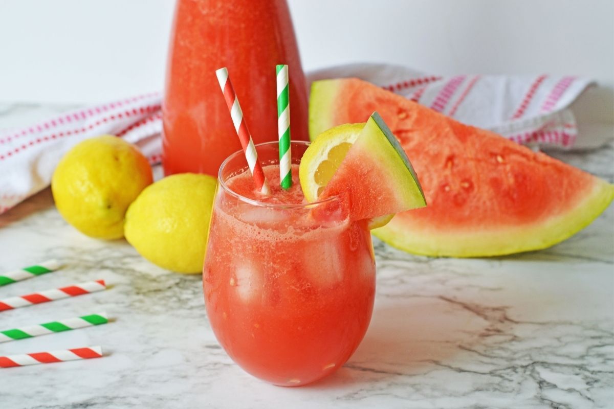 A hurricane glass on a table filled with red watermelon lemonade with two stripy straws in it with slices of watermelon and lemon on the rim of the glass. 
