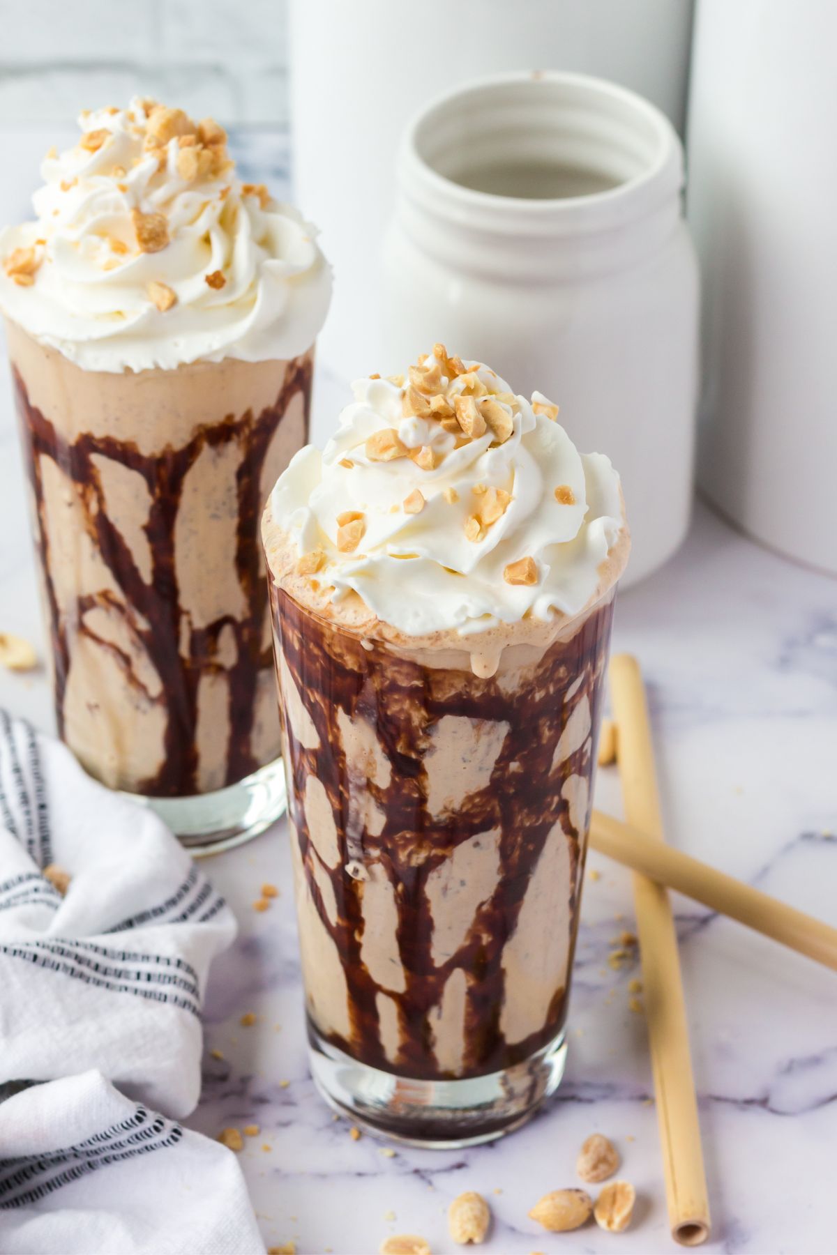 two milkshakes with chocolate sauce drizzled on the inside and topped with whipped cream and peanuts