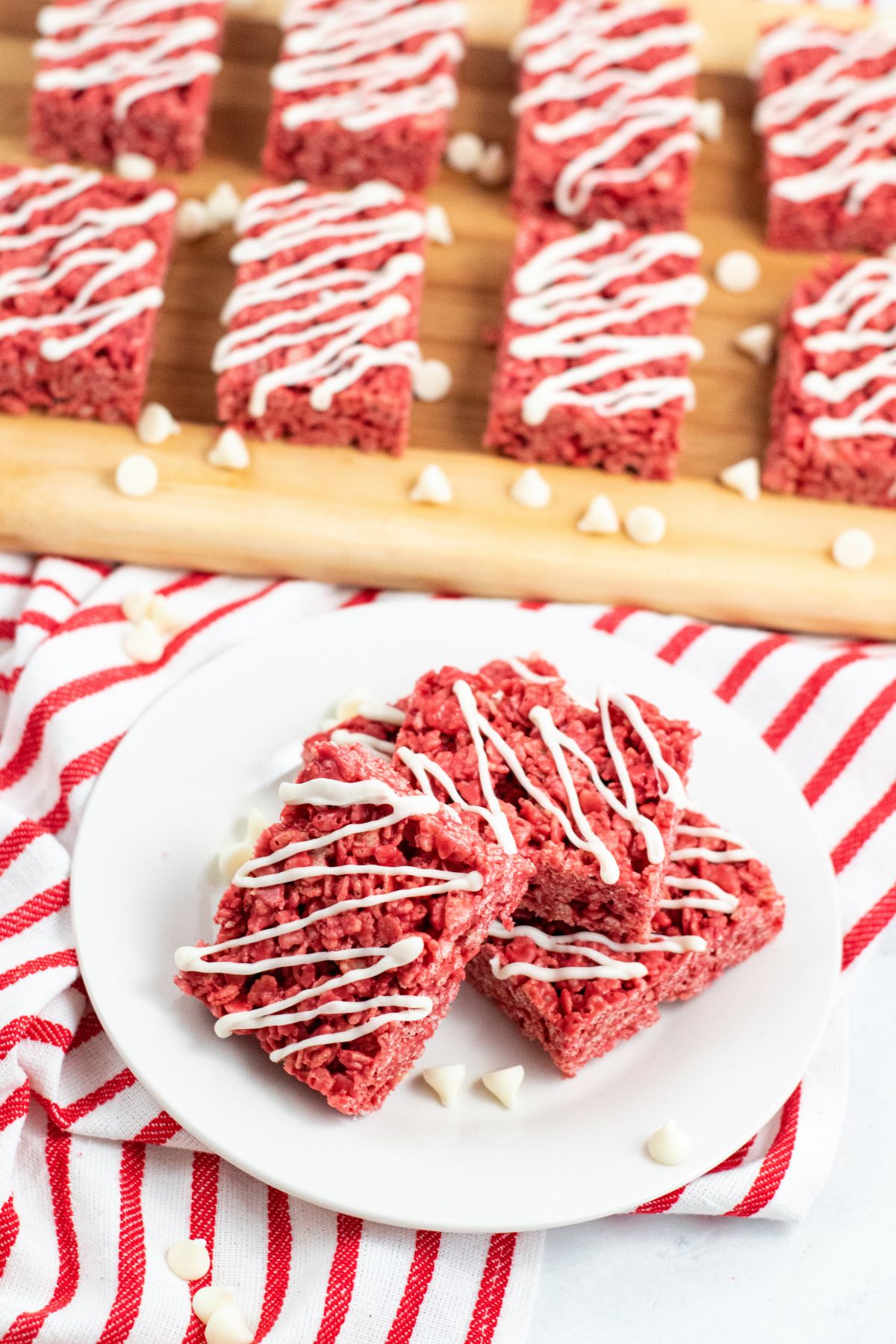 Rice Krispie Treats on a white plate with white chocolate drizzled on top on a table with a red and white stripy tea towel. 