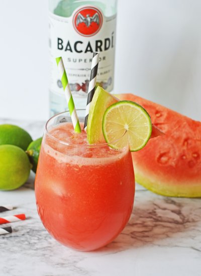 ALT Glass on a worktop with a pink watermelon rum punch in it with two stripy straws in the glass and a slice of lime and watermelon on the rim of the glass. There is a bottle of Bacardi rum in the background next to limes and slices of watermelon.