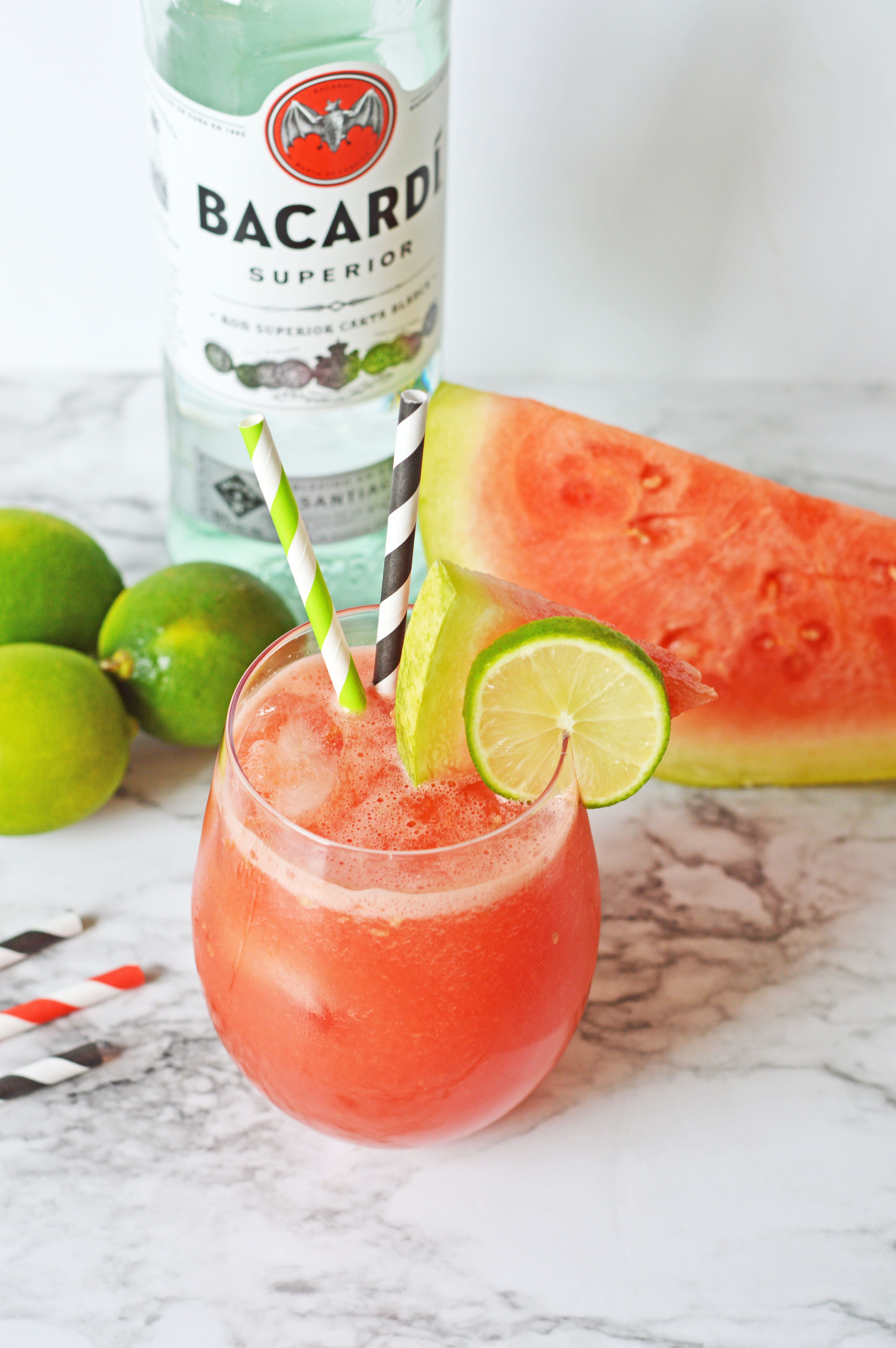 ALT Glass on a worktop with a pink watermelon rum punch in it with two stripy straws in the glass and a slice of lime and watermelon on the rim of the glass. There is a bottle of Bacardi rum in the background next to limes and slices of watermelon.