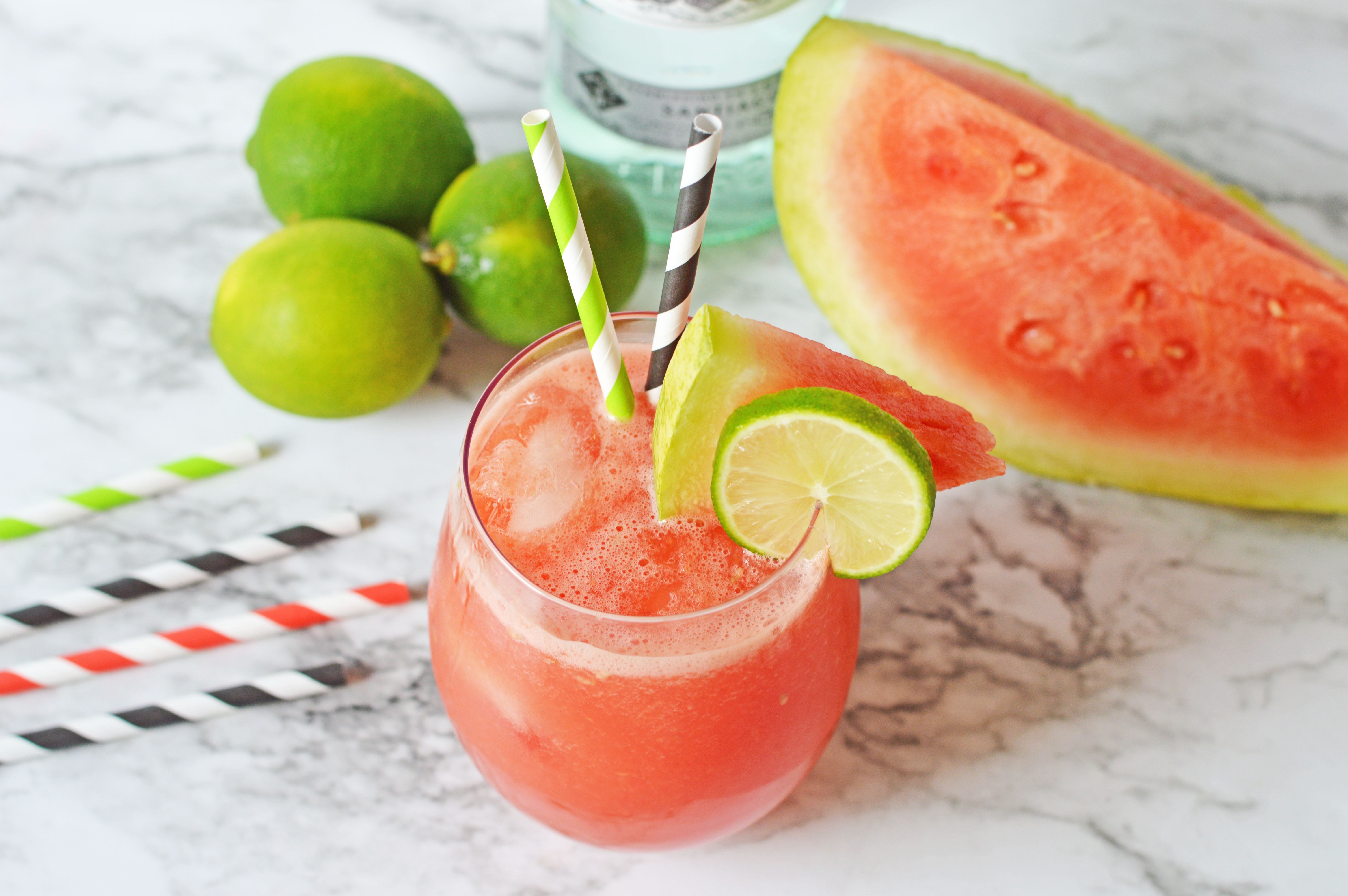 Glass on a worktop with a pink watermelon rum punch in it with two stripy straws in the glass and a slice of lime and watermelon on the rim of the glass. There is a bottle of Bacardi rum in the background next to limes and slices of watermelon.