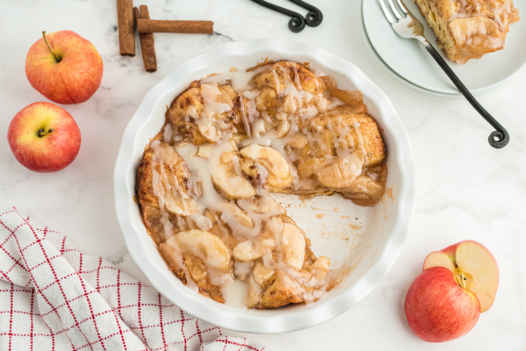 Top view of cinnamon Roll Apple Bake in a white pie dish with frosting drizzled over it and a slice taken out of it. There are whole and sliced apples to the side.of the dish as well. 