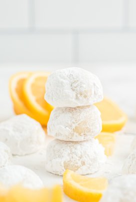 Three round cookies covered in powdered sugar stacked on top of each other in a tower on a worktop, surrounded by other round cookies covered in powdered sugar, with thick slices of lemon in the background.