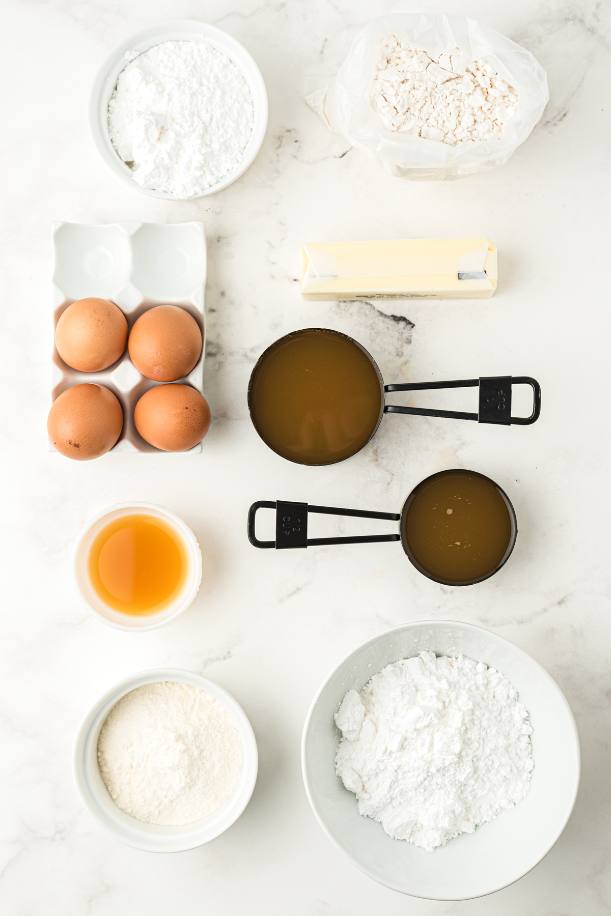 Ingredients for Apple Cider Cake on a white worktop in small bowls including white cake mix, pudding mix, eggs, butter, apple cider, powdered sugar, and confectioner's sugar. 