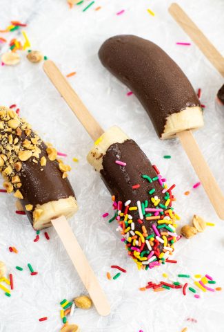 Top view close-up of three banana pops covered in chocolate lying on parchment paper, one plain, one with rainbow sprinkles on it and the last one with crushed peanuts on it.