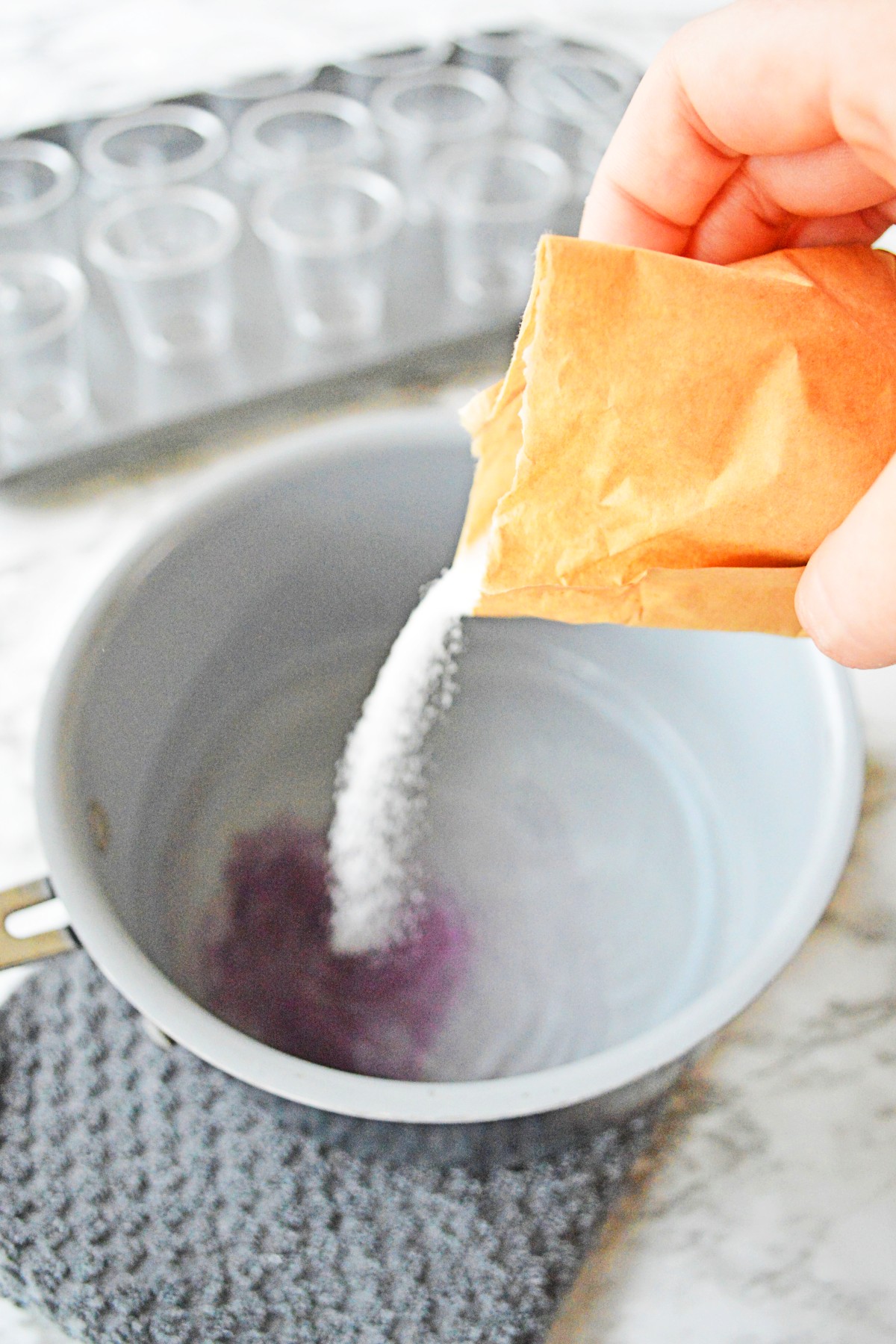 White bowl with water and vodka in it with a packet of jello powder being poured into it and the mixture is turning purple on the bottom. 