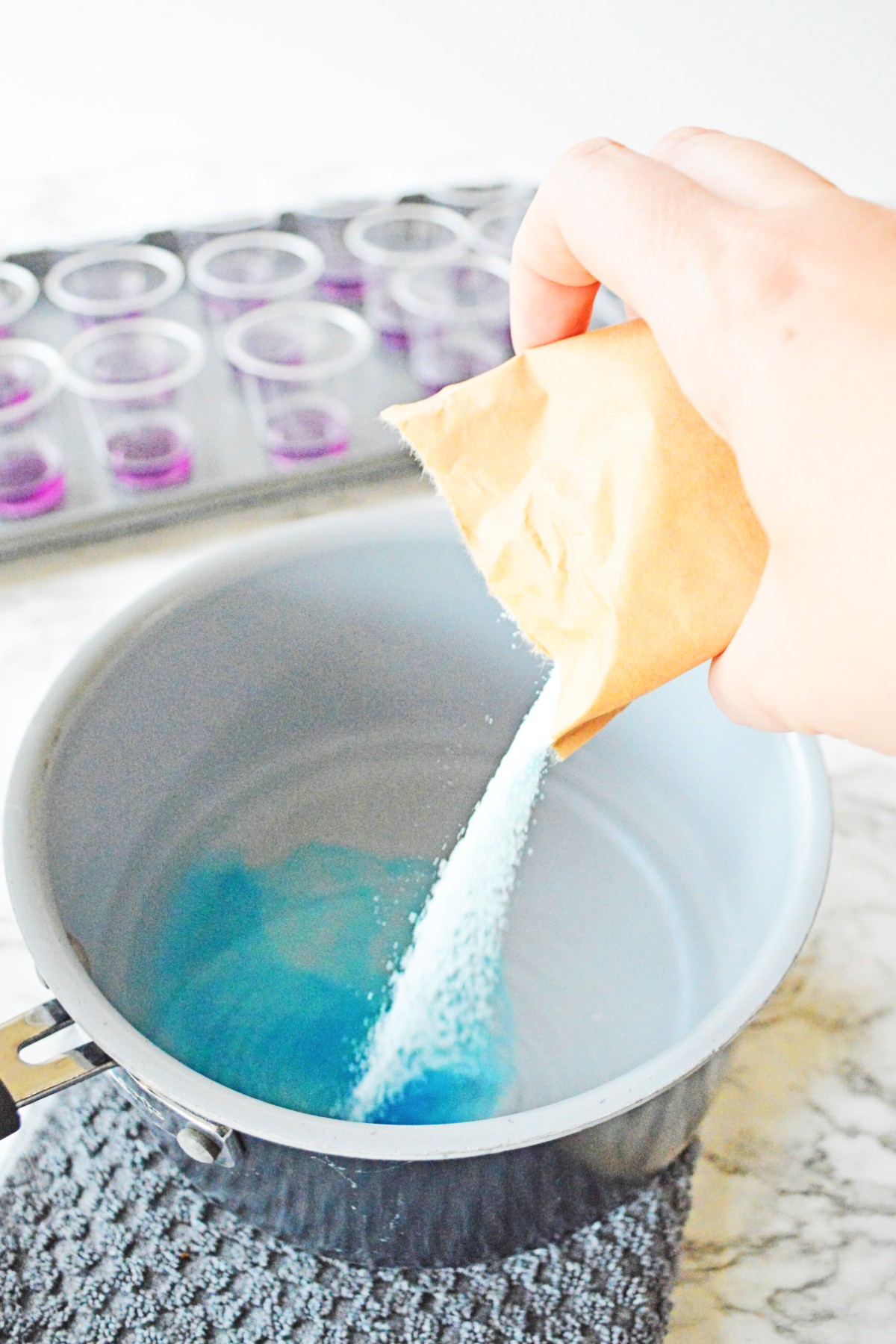 White bowl with water and vodka in it with a packet of jello powder being poured into it and the mixture is turning blue on the bottom.