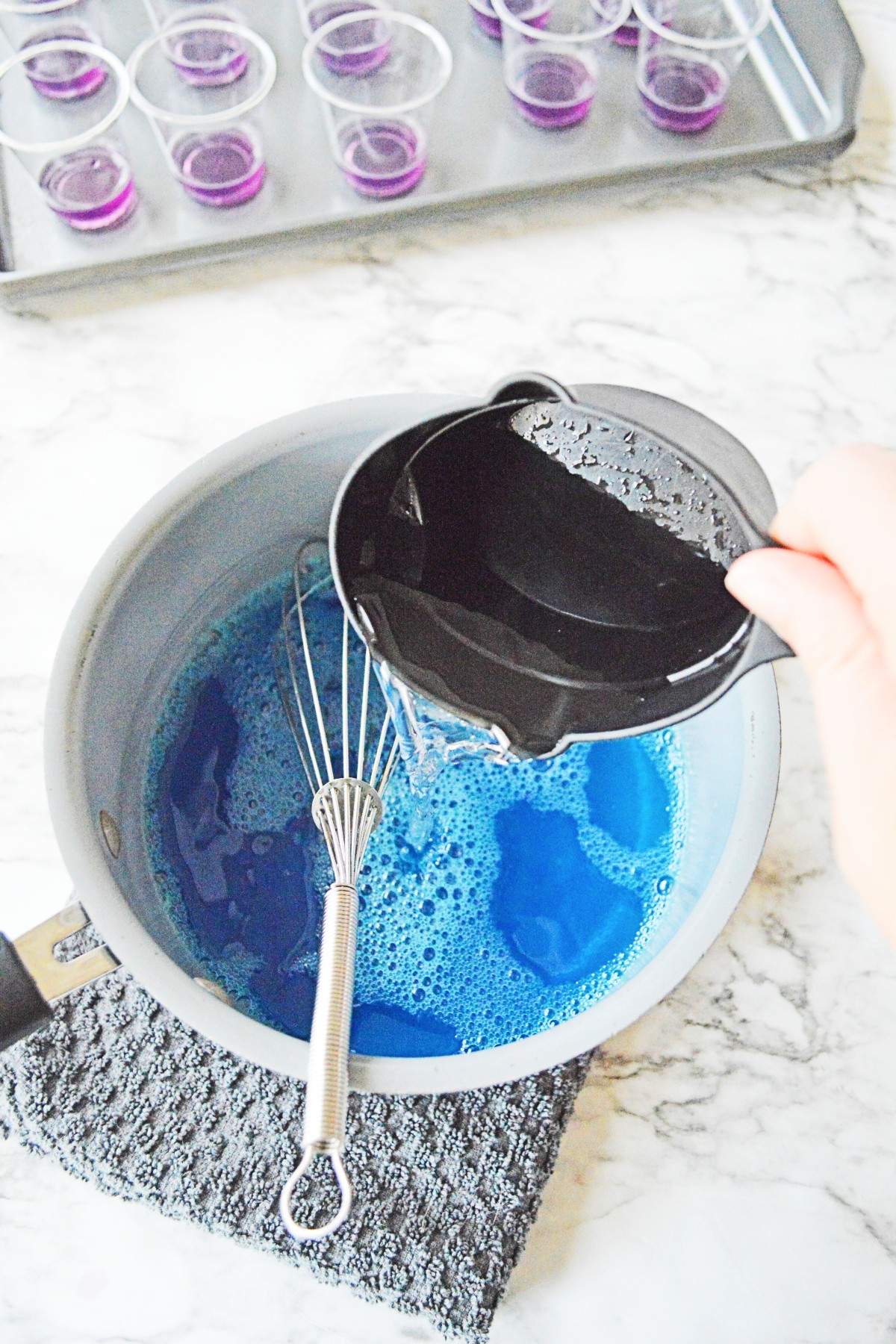 White bowl with a whisk and blue jello mixture in the bottom and a measuring cup with vodka being poured into it.