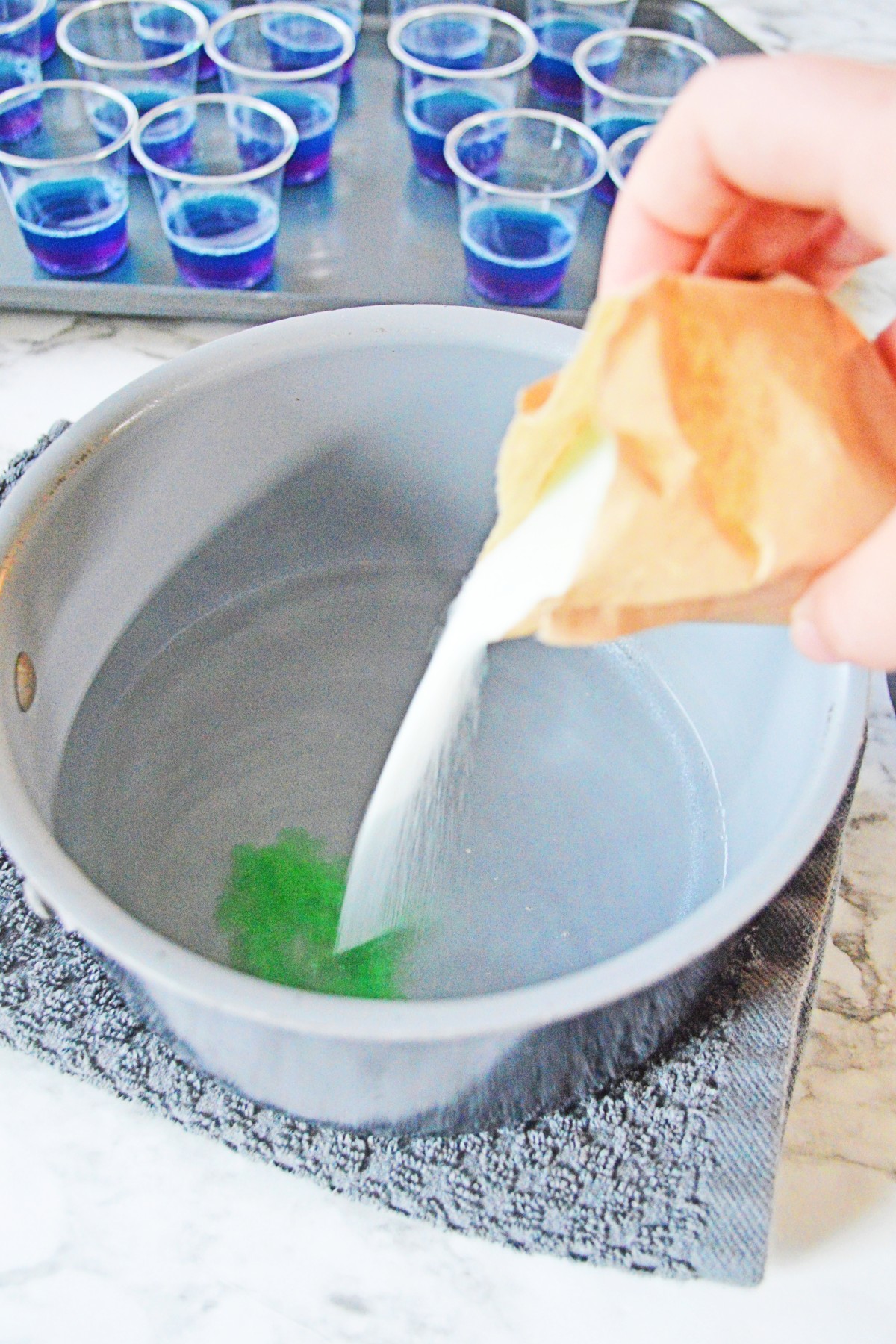 White bowl with water and vodka in it with a packet of jello powder being poured into it and the mixture is turning green on the bottom.