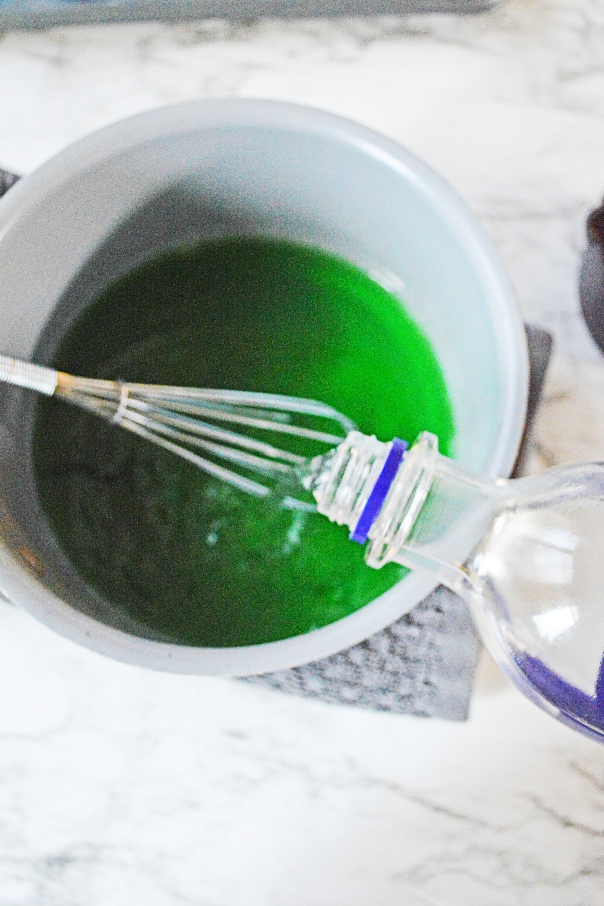 White bowl with a whisk and green jello mixture in the bottom and a measuring cup with vodka being poured into it.