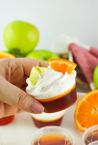 A sangria shot in a small plastic condiment cup with whipped cream and small slices of apple and orange on top, being held in mid air by a hand.