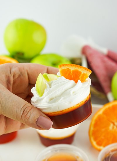 A sangria shot in a small plastic condiment cup with whipped cream and small slices of apple and orange on top, being held in mid air by a hand.