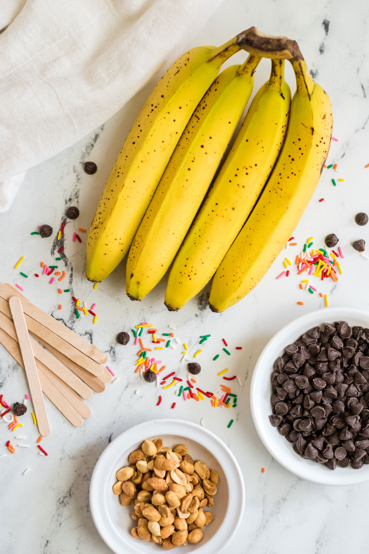 Ingredients of Banana Pops on a white counter including bananas, chocolate chips, popsicle sticks, chopped peanuts and sprinkles. 
