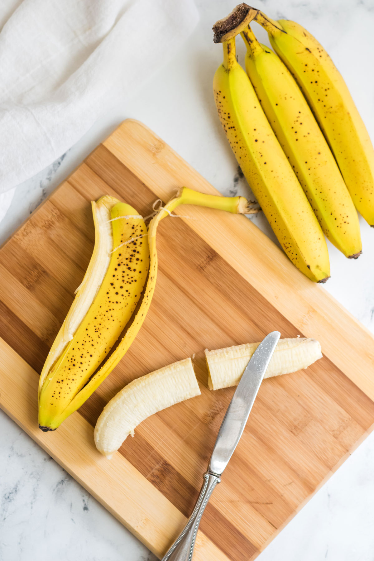 Top view of a chopping board with a banana cut in half on it, and a bunch of whole bananas sitting next to the board. 