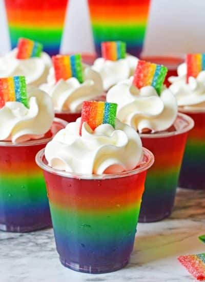 A marble worktop with several cups filled with layered rainbow jelly with cream and a rainbow candy on top.