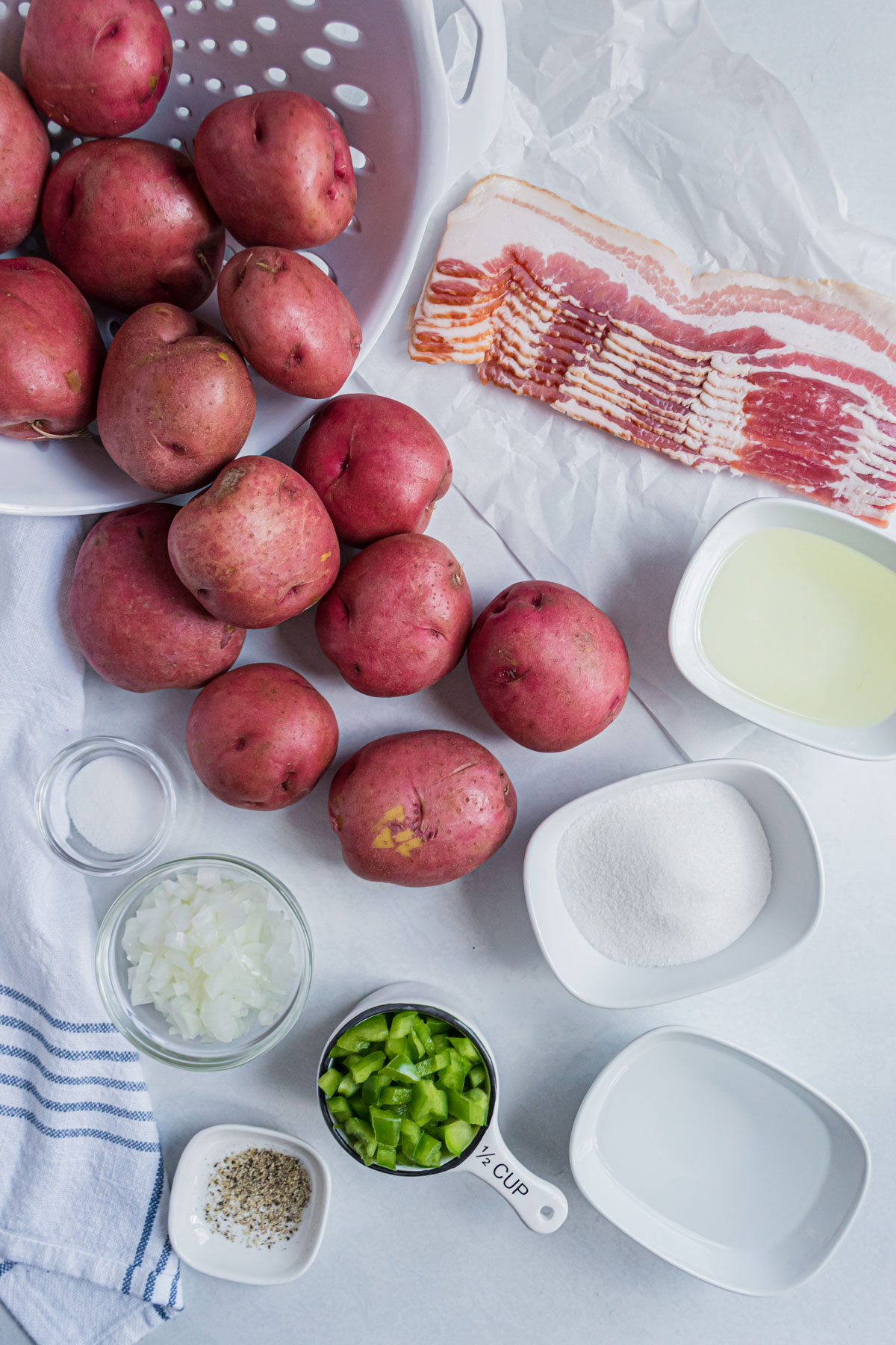Flatlay of ingredients of Hot German Potato Salad. This includes small red potatoes, bacon, onion, green pepper, salt, black pepper, white vinegar, tablespoons of sugar and vegetable oil.