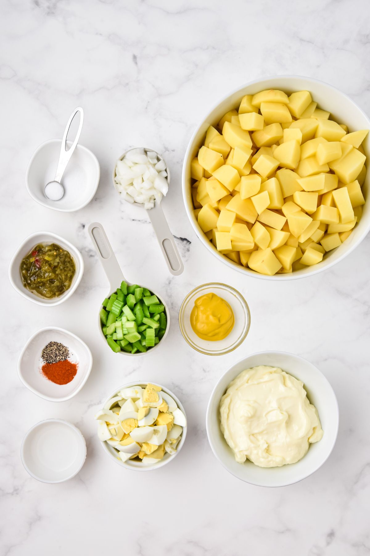 ingredients on white counter: diced potatoes, mustard, mayonnaise, chopped celery, chopped hard boiled eggs, relish, vinegar, and seasings