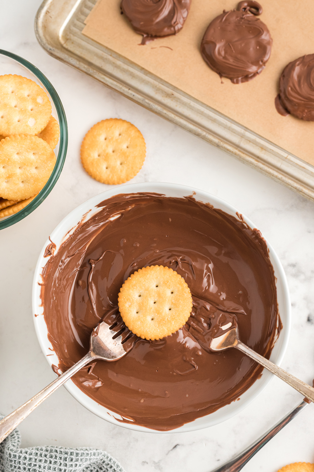 White mixing bowl filled with melted chocolate with two forks and a ritz cracker in the middle of it. This is one step in preparing Ritz Cracker Thin Mints