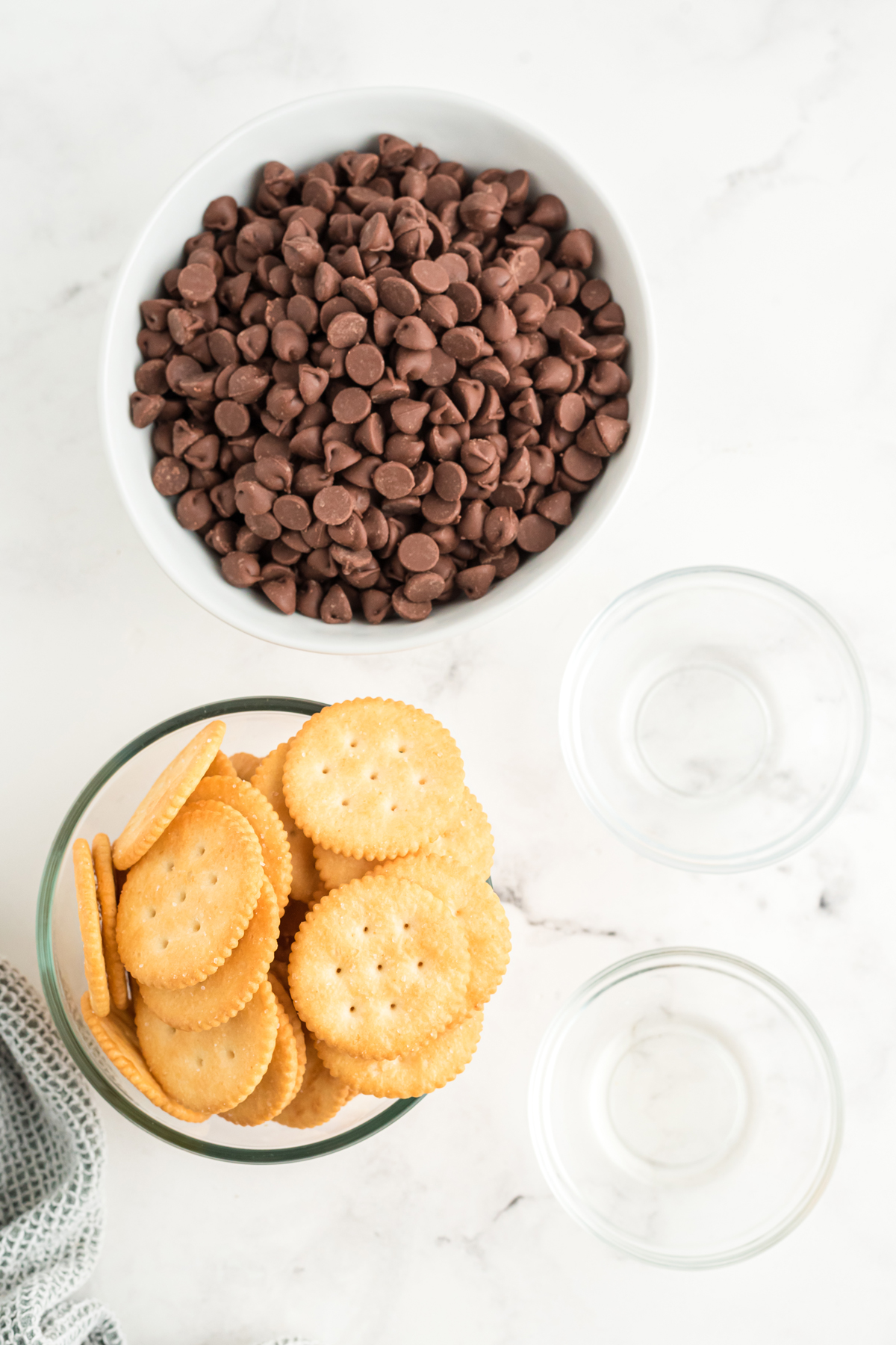 Top view of a white bowl filled with chocolate chips, a glass bowl filled with Ritz crackers, and two small glass bowls one with oil in it and the other with Peppermint extract in it.  This is for Ritz Cracker Thin Mints recipe