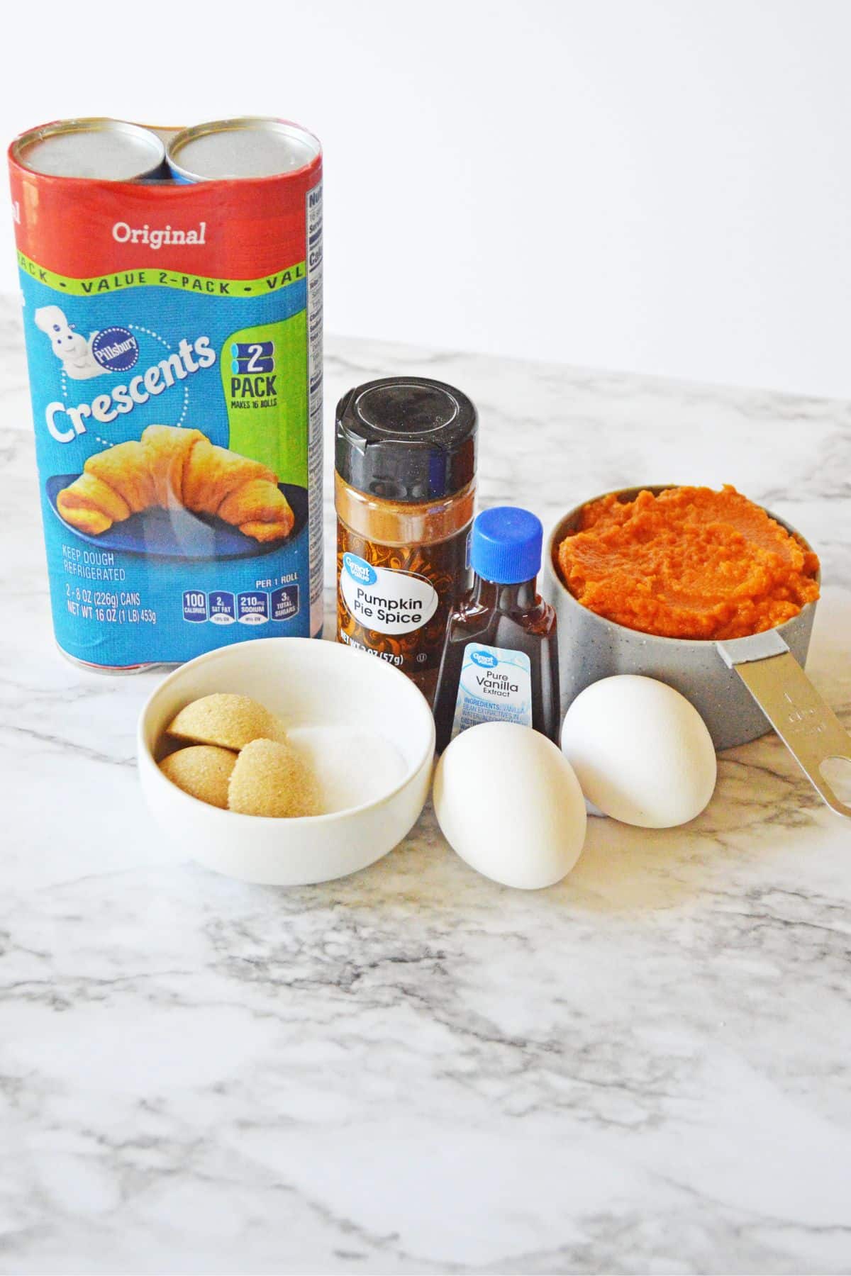 ingredients on marble counter: crescent roll packs, pumpkin pie spice, vanilla, 2 white eggs, pumpkin puree, brown and white sugar